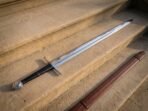 Steel disc pommel sword with black leather grip and straight cross guard on sandstone steps beside a brown leather scabbard