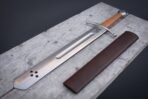 Steel falchion with curved guard, tan leather grip and faceted pommel on grey planks beside a brown leather scabbard