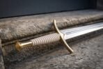 Steel fantasy arming sword with engraved brass cross guard, ribbed pale grip and brass pommel resting on a stone step