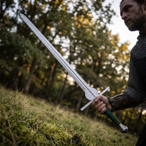 Steel feder training sword with a rounded schilt and green cord grip held low by a man in mail with a leather bracer on a grassy bank