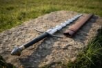 Steel flamberge sword with faceted cone pommel and black grip on a flat rock in grass with its brown leather scabbard
