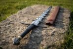 Steel flamberge sword with wavy blade, blackened hooked guard and black grip lying on a rock beside a brown scabbard