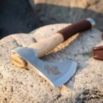 Steel hand axe head close up with etched Norse raven, polished cheek and ash haft resting on pale rock