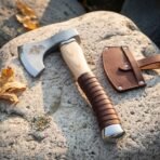 Steel hand axe with etched raven head, pale ash haft and wrapped leather grip on a stone beside its leather sheath