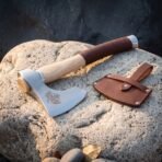 Steel hand axe with raven etch and pale ash haft on a boulder beside an open brown leather sheath with snap