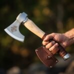 Steel hand axe with raven etch held by the leather wrapped grip, brown snap sheath hanging from the hand