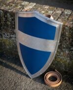 Steel heater shield with blue painted face and engraved Celtic knot band leaning on a brick wall beside a leather strap