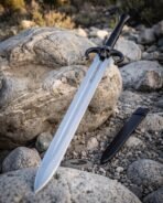 Steel horned skull sword with blackened ram horn guard and black grip propped against a boulder on a rocky shore, scabbard below