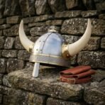 Steel horned Viking helmet with nasal bar and brass horn sockets on a stone ledge beside a brown leather strap