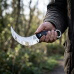 Steel karambit knife with curved polished blade, black micarta scales and finger ring held in a hand on a woodland path