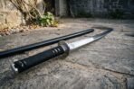 Steel katana with black cord wrapped tsuka and polished tsuba laid on flagstones with its black saya, close up from the hilt end