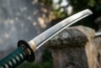 Steel katana blade with a visible hamon, green ito wrapped tsuka and brass habaki, laid across a stone pillar in a garden