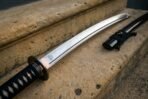 Steel katana close up of the dark tsuba, copper habaki and Valknut mark on the blade over a black cord handle