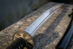 Steel katana close up of the hamon line and dark round tsuba engraved with a gold runic compass, on stone