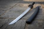 Steel katana with etched knotwork band and blackened tsuba laid flat on grey paving stones with its black saya beside it