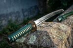 Steel katana with green ito wrapped tsuka and iron tsuba laid on a lichen covered stone ledge with the green lacquered saya behind