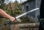 Steel katana with a green wrapped tsuka held out in one hand over a stone gate post, green lacquered saya at the waist
