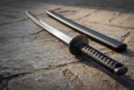 Close up of a steel katana hilt with blackened tsuba and black cord wrapped tsuka on flagstones, black saya lying behind
