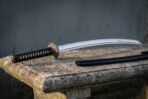 Steel katana in side view on a weathered stone bench with black wrapped handle and black lacquered saya behind