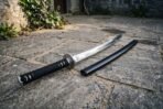 Steel katana with polished tsuba and rayskin under black cord wrap lying on a stone courtyard floor next to its black saya