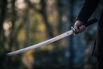 Steel katana with valknut etched blade held out in one hand in dark woodland, black saya held in the other hand behind