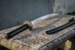 Steel katana with black cord wrapped handle and engraved tsuba lying on a stone bench beside its black saya