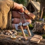 Steel keyring with miniature axe and hammer hanging from a finger above a campsite with a tent and pack behind