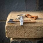 Steel keyring with miniature axe and hammer laid on a sandstone block beside its tan leather snap tab