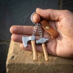 Steel keyring with a miniature axe and valknut hammer on wooden hafts resting in an open palm with a leather tab
