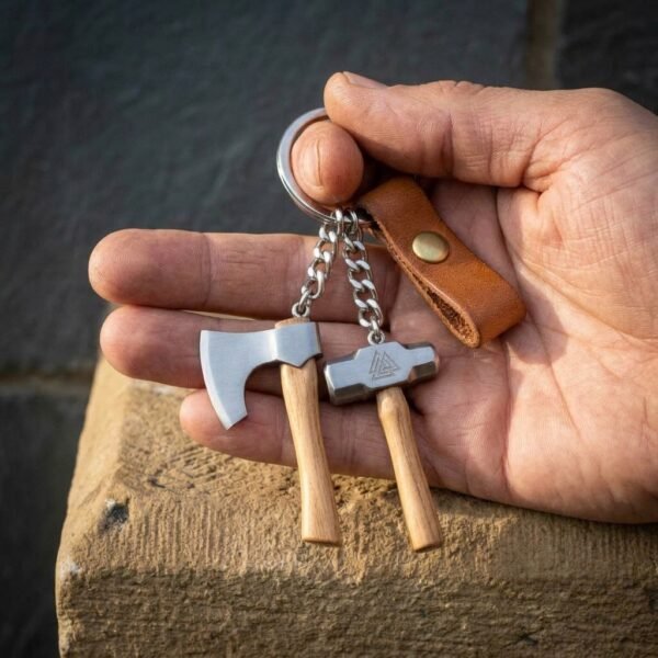 Steel keyring with a miniature axe and valknut hammer on wooden hafts resting in an open palm with a leather tab