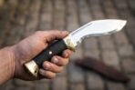 Steel kukri knife with a black cord wrapped grip, raven engraved brass bolster and flared brass butt cap held in hand over cobblestones, sheath behind