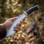 Steel kukri knife with a black horn handle and brass bolster held in one hand in the woods, a line of runes etched along the polished blade