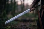 Steel langseax with knotwork etched blade and leather wrapped grip held low in a gloved hand in a dark forest