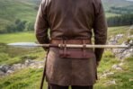Steel leaf blade spear carried across the back of a man in a leather jerkin, held by belt loops on a green hillside