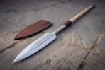 Steel leaf blade spear head close up with Elder Futhark runes, red leather wrap and brown sheath on flagstones