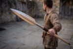 Steel leaf blade spear with rune row held forward in one hand by a man in a stone yard, head close to the camera