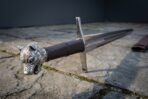 Close up of a steel longsword hilt with cast wolf head pommel, brown leather grip and etched knotwork ricasso on paving slabs
