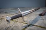 Steel longsword with wolf head pommel, long leather grip and straight cross guard laid on stone paving, scabbard tip at the edge