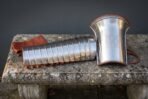 Steel manica arm guard with brass riveted bands and russet leather straps next to an upright flared knotwork bracer on a lichen covered stone bench