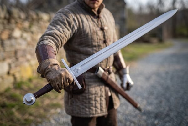 Steel medieval arming sword with a wheel pommel held in a gloved hand by a man in a padded gambeson, brown leather scabbard at his belt