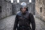 Steel nasal helmet with a riveted dome and brow band worn by a bearded man in a dark tunic inside a stone castle courtyard