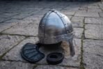 Steel nasal helmet with riveted seams and a brow band on stone flags beside its padded black liner and leather strap