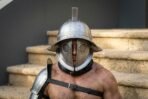 Steel Roman helmet with round pierced eye grilles, wide brim and crest fin worn by a man with a steel shoulder guard and black leather straps