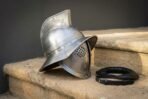 Steel Roman helmet with tall crest fin, wide brim, Greek key brow band and round pierced eye grilles, side view on sandstone steps