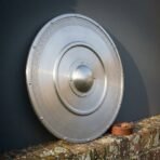Steel round shield with riveted rim and engraved knotwork border leaning on a brick wall beside a coiled leather arm strap
