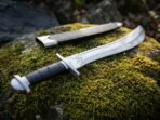 Steel sawback short sword with chevron mark at the ricasso and black wrapped grip laid on moss with its black scabbard