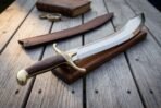 Steel scimitar resting on a wooden display block with its brown leather scabbard, an old book and a compass behind