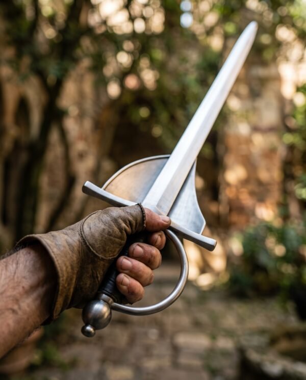 Steel shell guard rapier trainer held in a gloved fist with the blade angled up in a sunny garden