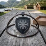 Steel shield pendant with blackened helm of awe stave and knotwork edge, brown leather cord looped on grey timber, wooden box behind