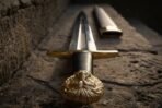Close up of a steel short sword with cast brass lion head pommel, black grip and brass guard on a stone ledge, scabbard behind